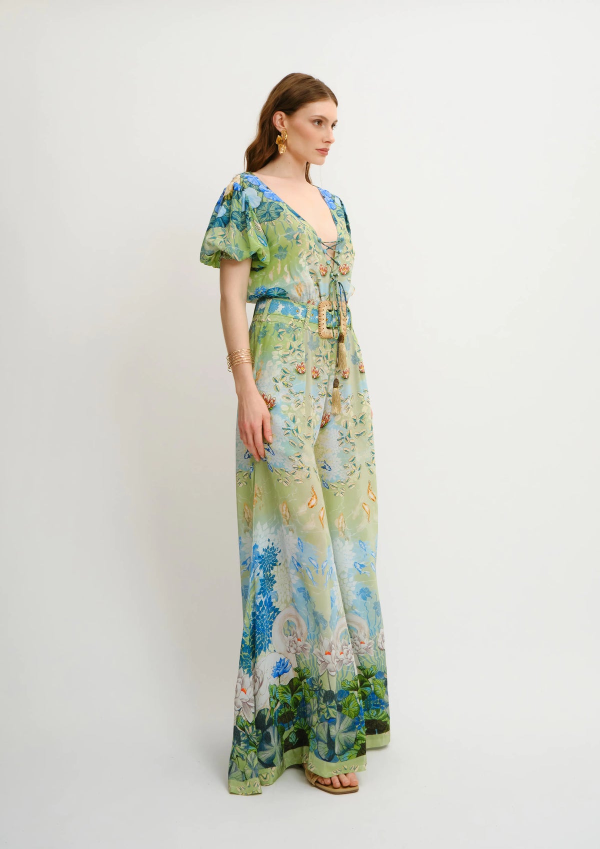 A woman models a long-sleeved Koré Collections maxi jumpsuit with a vibrant, painterly floral and landscape print in shades of green, blue, and yellow, set against a plain light gray studio background.