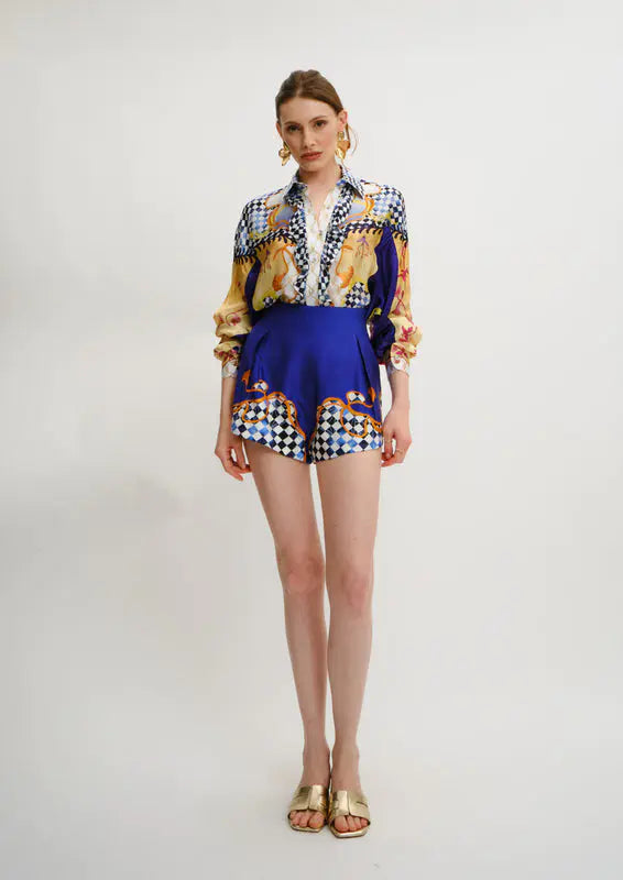 A model stands against a light gray studio background wearing a Koré Collections matching two-piece outfit with a long-sleeve button-up shirt and shorts. The top features a multicolor abstract print with blue, yellow, black, and white, while the shorts are solid royal blue with a checkered hem.