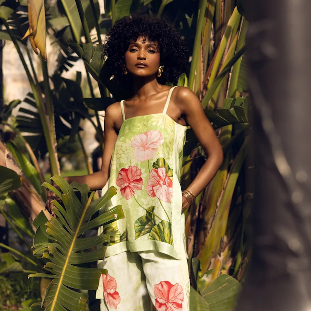 A woman wearing a light green sleeveless jumpsuit with a pink hibiscus floral print, standing in a tropical garden with large green leaves.