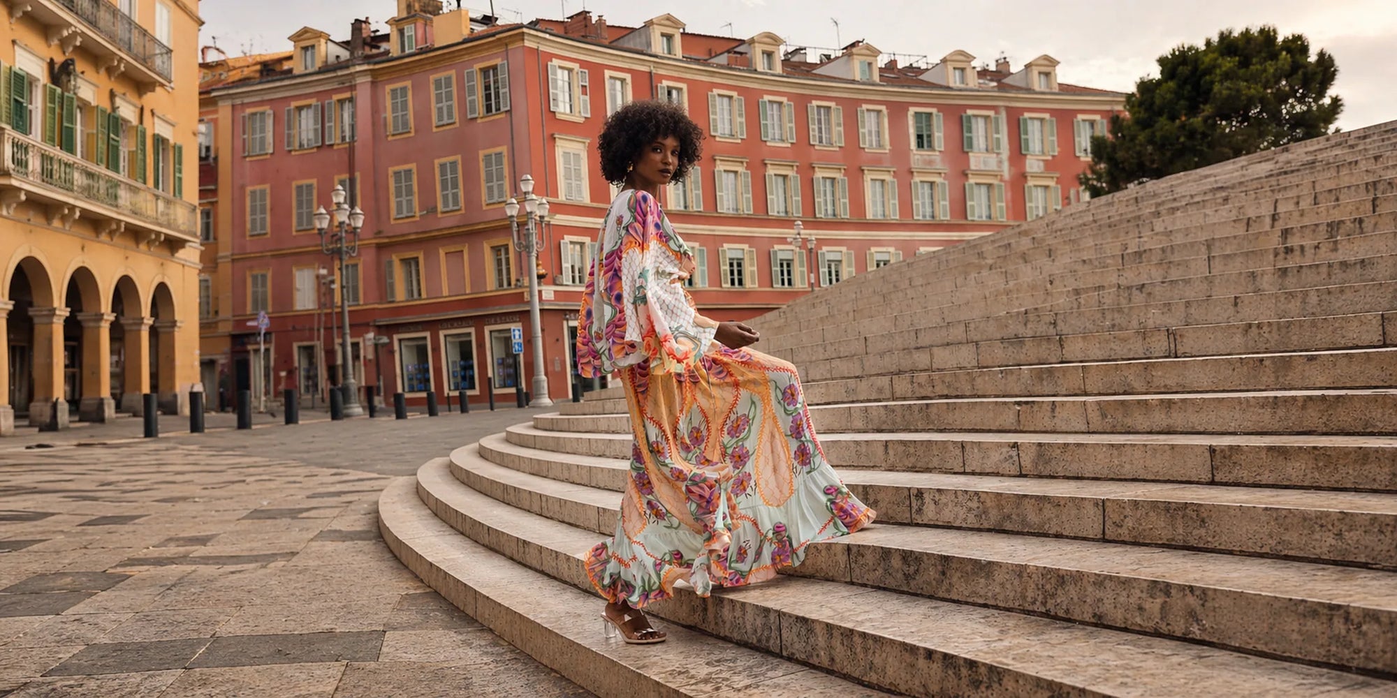 A woman with an afro hairstyle wearing a long, colorful floral print maxi dress walks down wide stone steps in an outdoor European-style plaza with orange and yellow buildings in the background.