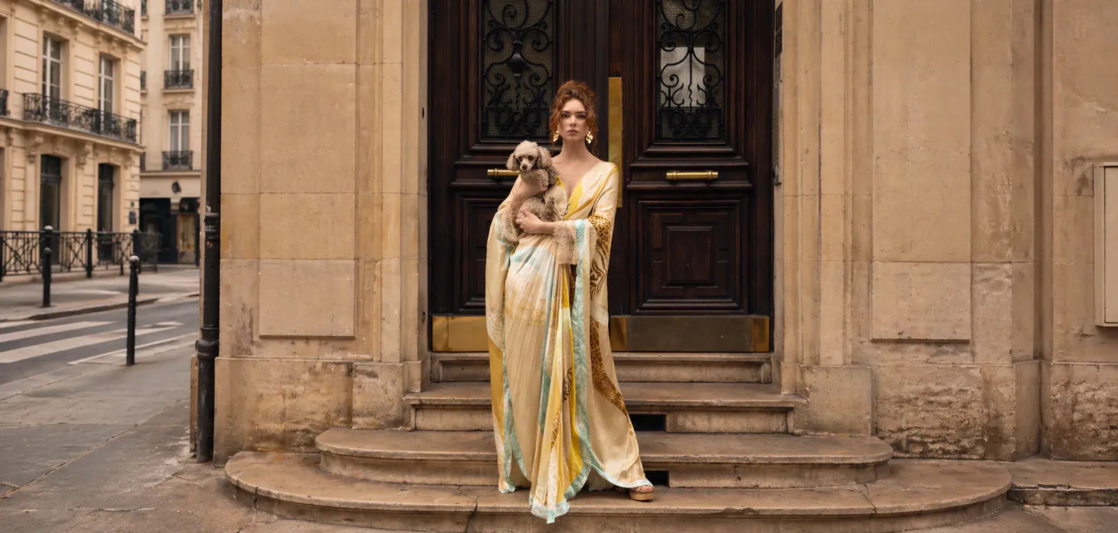 A woman stands on stone steps in front of a beige building, holding a small dog. She is wearing a flowing, draped satin gown in pale yellow, cream, and light blue tones.