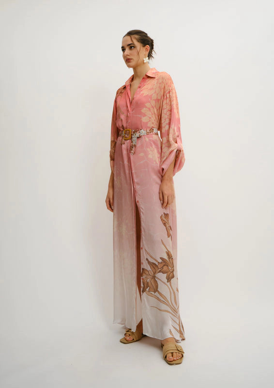 A woman models a long-sleeved, wide-leg koré collections maxi shirt dress in a dusty pink color with a brown floral pattern and a matching belt, standing against a plain white studio background.