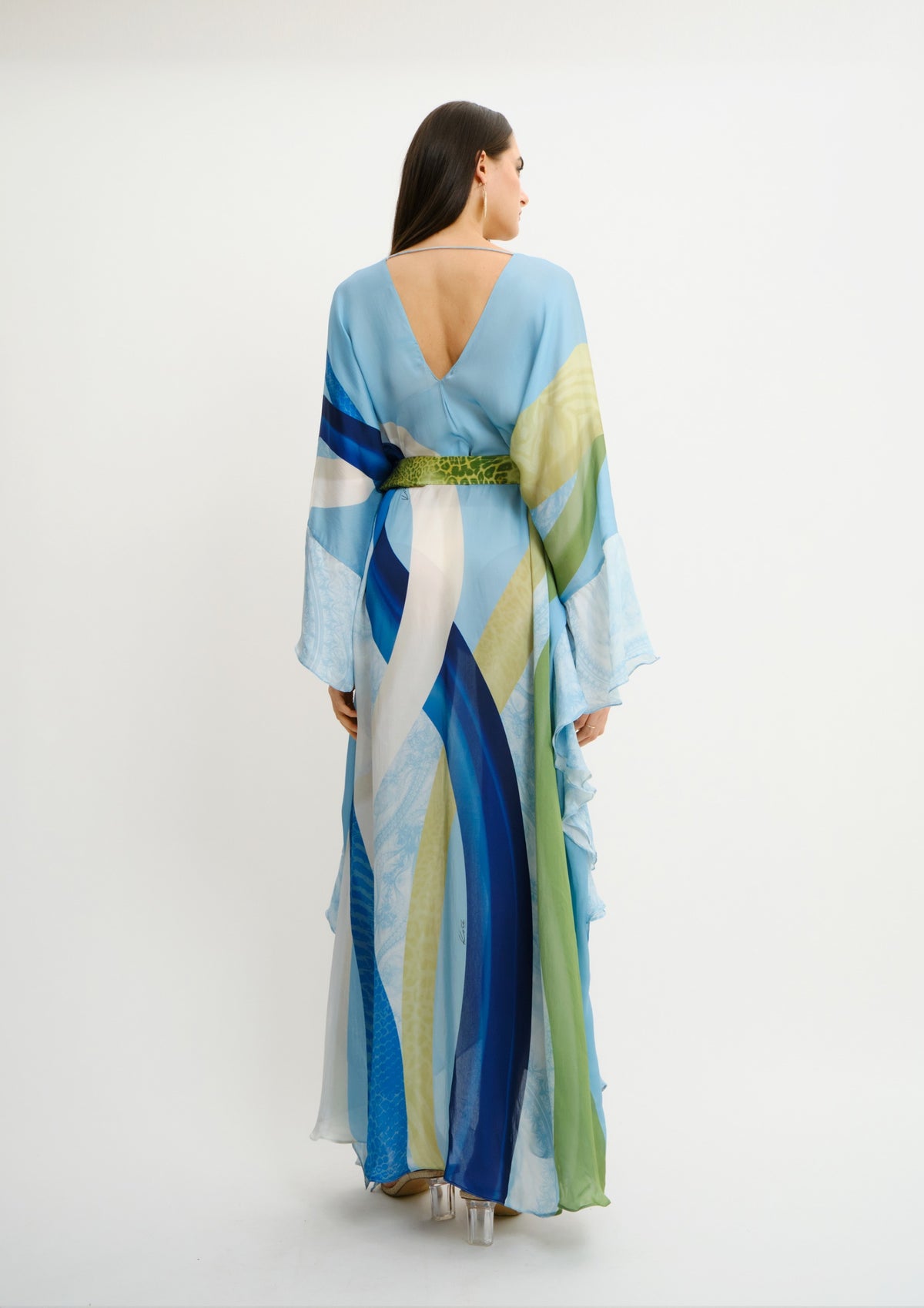 A woman viewed from the back wearing a flowing Koré collections maxi dress with an abstract watercolor-like print in light blue, navy, white, and green, featuring a deep V-neckline and a green textured waistband, set against a plain white studio background.