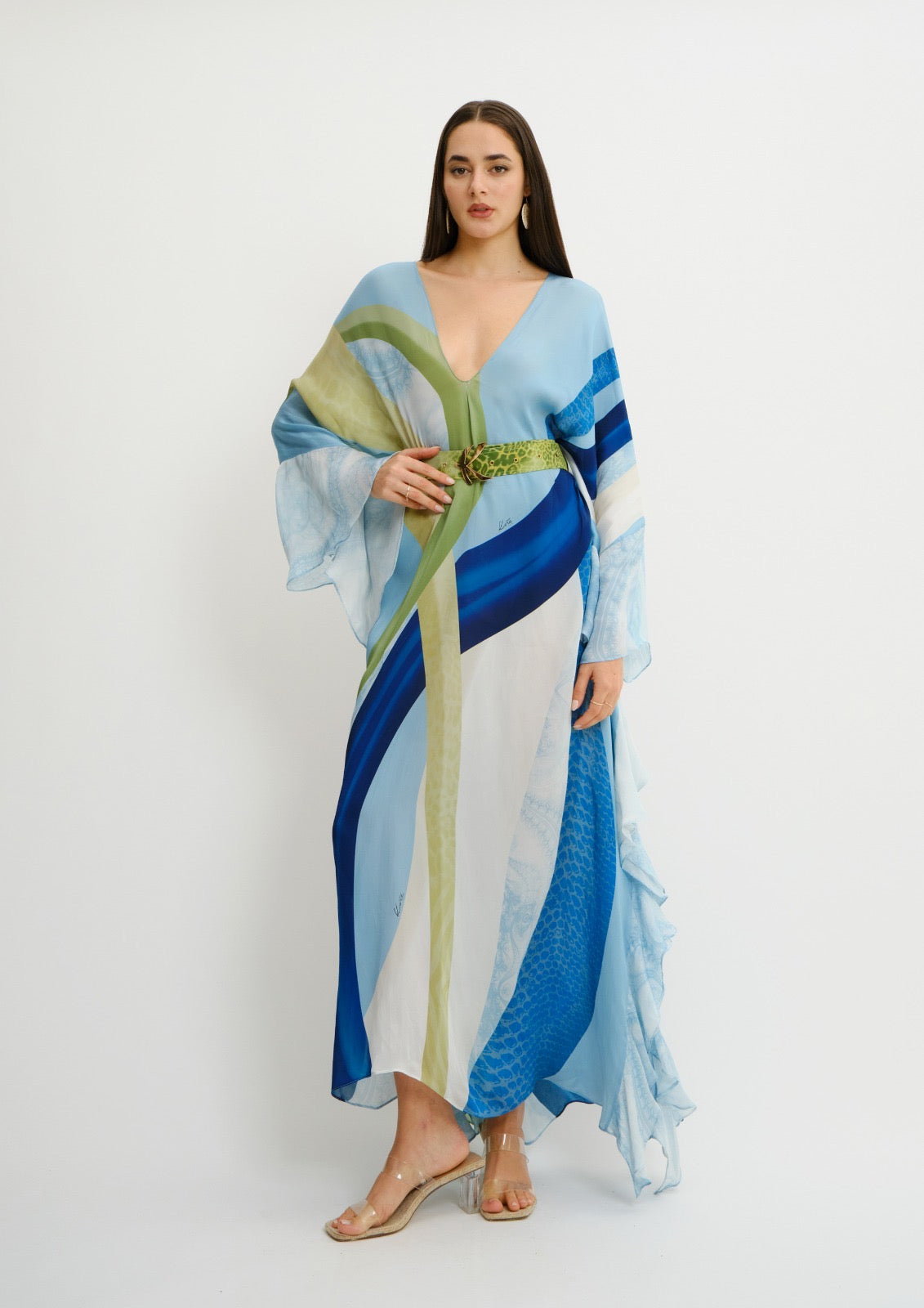 A woman modeling a long, flowing koré collections kaftan dress with abstract blue, white, and olive green stripes, styled with a matching green belt and clear wedge sandals against a plain white studio background.