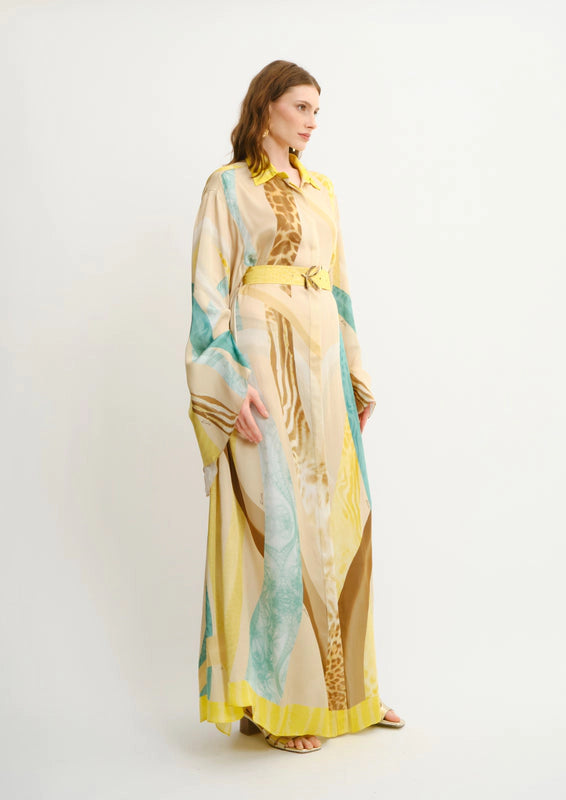 A woman modeling a long, flowing koré collections maxi shirt dress dress with a giraffe print pattern in beige, yellow, and brown, featuring turquoise blue side panels and a matching yellow belt, set against a plain white studio background.
