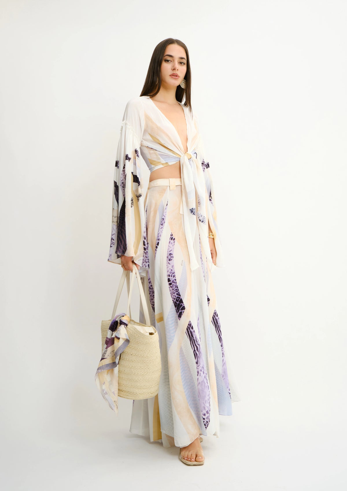 A woman modeling a matching Koré Collections two-piece bohemian print set consisting of a sheer long-sleeve crop top and a maxi skirt, holding a woven straw tote bag with a matching scarf against a plain white studio background.